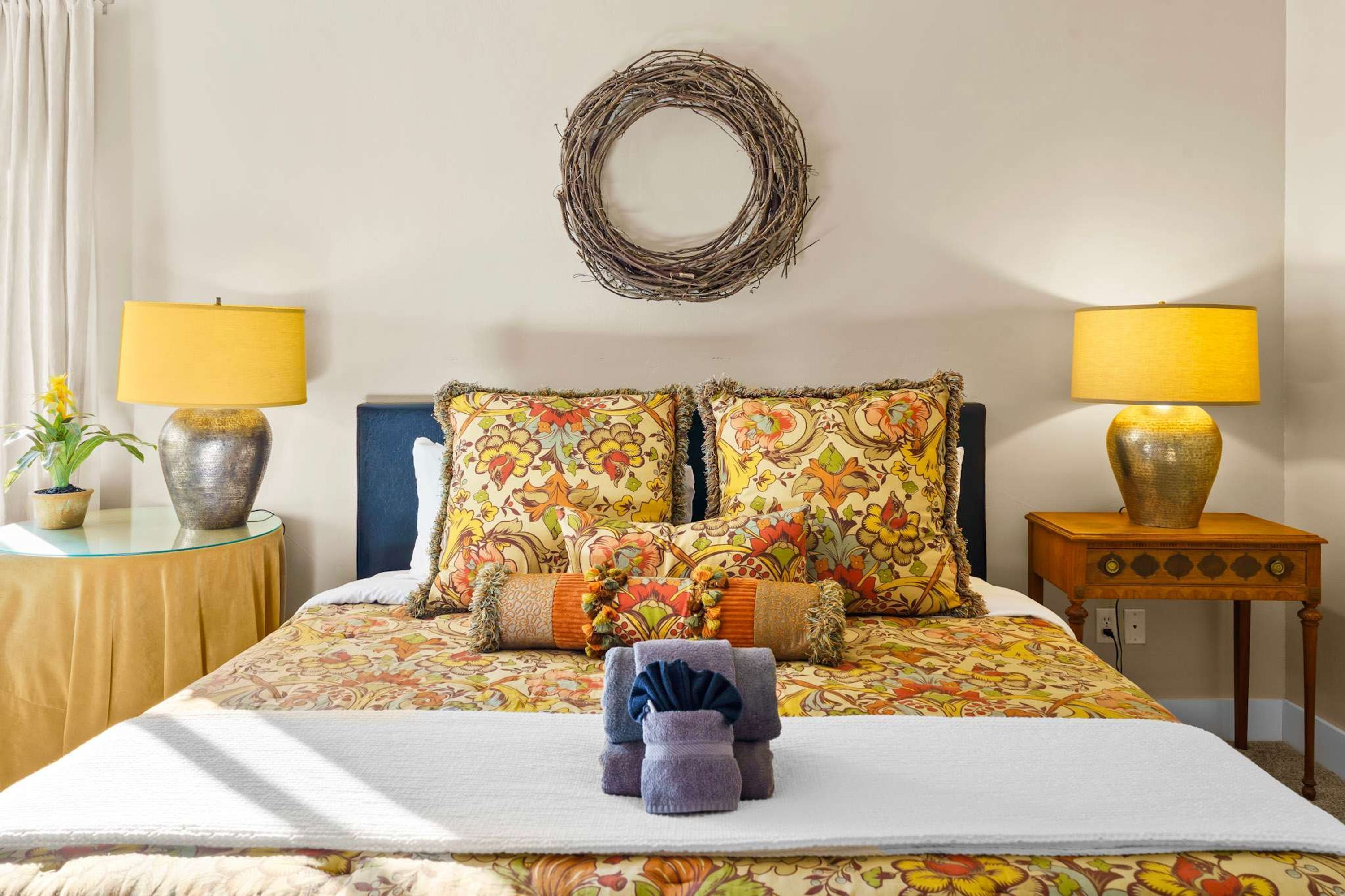 A neatly made bed with floral bedding, two bedside lamps, a wall-mounted circular mirror, and a small potted plant on the nightstand.