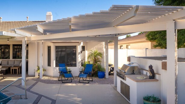 A covered outdoor seating area with two blue chairs, a barbecue grill, and a potted plant. - Pool Spa Outdoor Fireplace BBQ Pool Table (Goodyear)