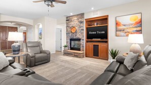 A modern living room with a stone fireplace, leather sofas, a flat-screen TV, and a ceiling fan. - Pool Spa Outdoor Fireplace BBQ Pool Table (Goodyear)
