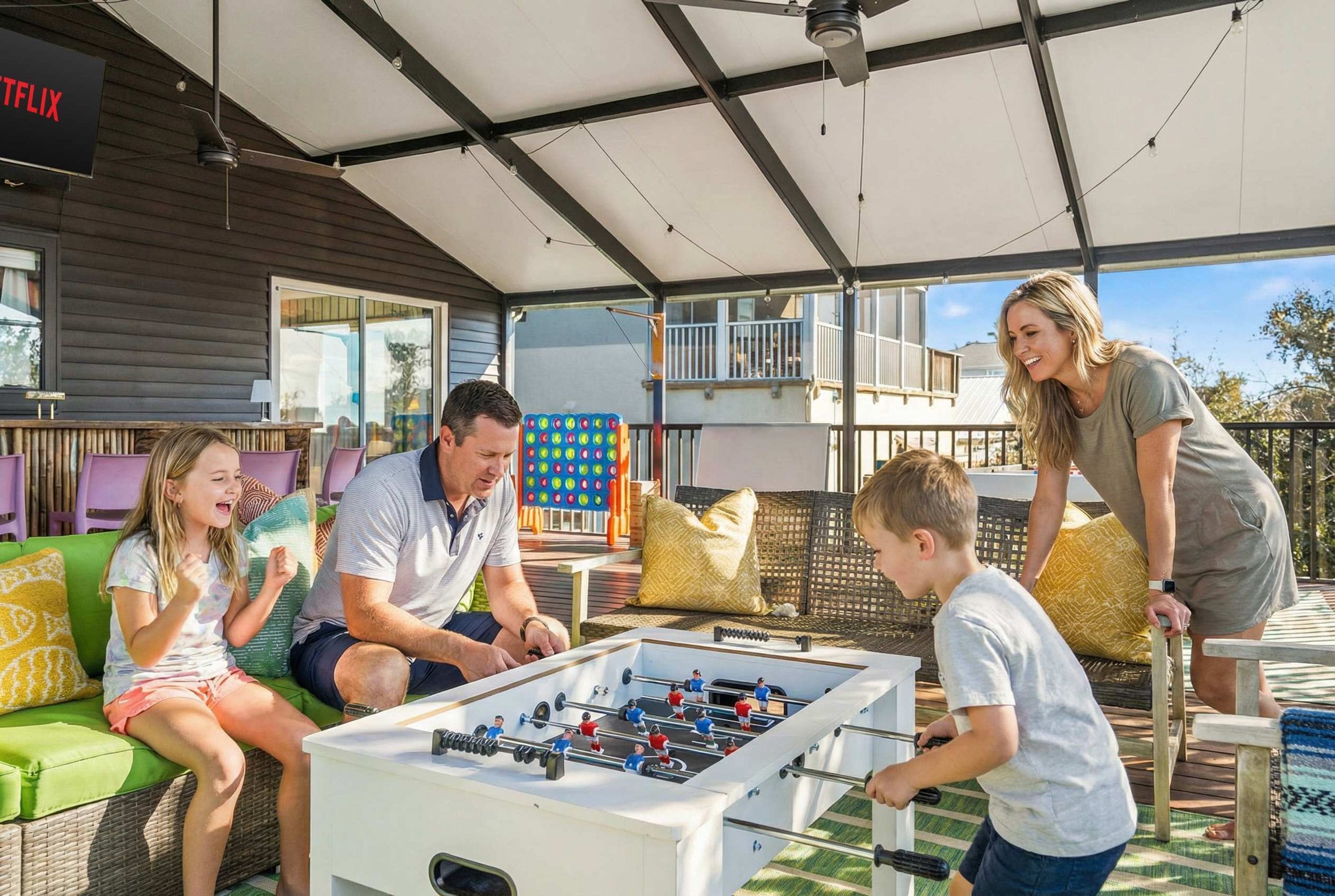 Eine Familie spielt Tischfußball auf einer überdachten Terrasse mit einer Couch und einem Tisch.