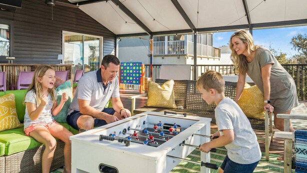 Une famille joue au baby-foot sur une terrasse extérieure dotée d’un espace abrité, d’un canapé et d’une table.