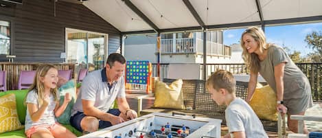 Une famille joue au baby-foot sur une terrasse extérieure dotée d’un espace abrité, d’un canapé et d’une table.