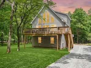 A two-story wooden house with a balcony, surrounded by trees and a gravel driveway.