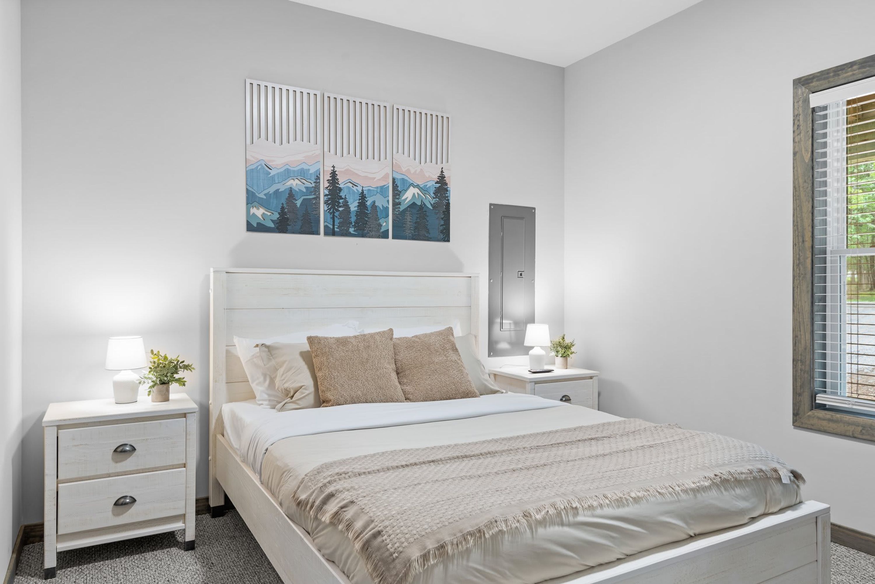 Une chambre à coucher comprenant un lit, des tables de chevet, une commode, une œuvre d’art accrochée au mur et une fenêtre avec des stores.
