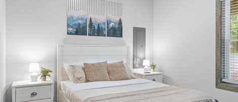 A bedroom with a bed, bedside tables, a nightstand, a wall-mounted artwork, and a window with blinds.