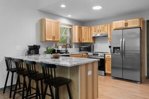 A modern kitchen with wooden cabinets, a stainless steel refrigerator, a built-in oven, and a bar area with stools.