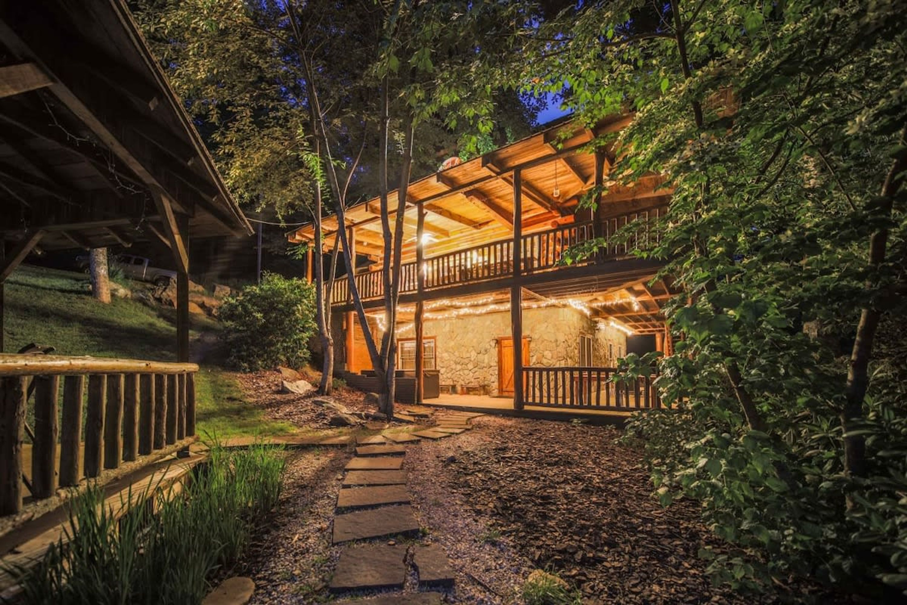 A two-story wooden cabin with a stone foundation, surrounded by trees and a stone pathway leading to the entrance.