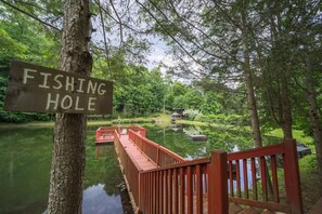 A wooden sign reading 'FISHING HOLE' attached to a tree, a wooden bridge over a calm body of water, and a gazebo in the background. - Lakeside Cabin - 2 Fireplaces-hot Tub Sauna Games (Mars Hill)