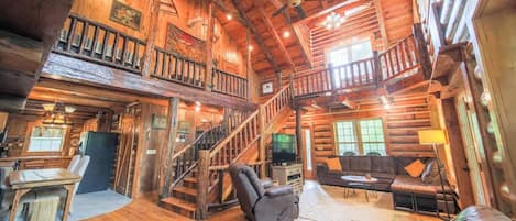 A spacious log cabin interior with a central staircase, a cozy seating area, and a television.