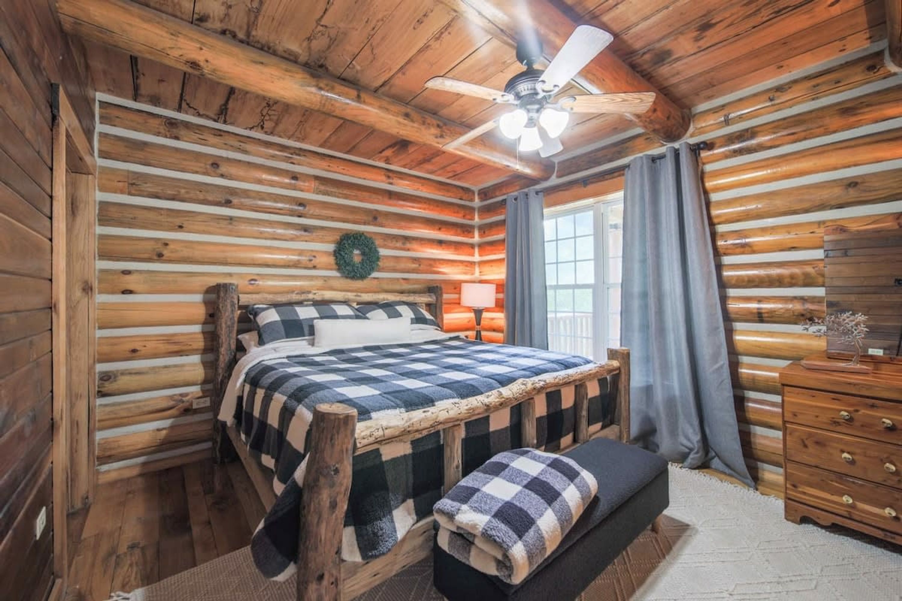A log cabin bedroom with a bed, bench, dresser, and a ceiling fan.