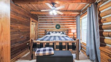 A log cabin bedroom with a bed, a ceiling fan, a window with curtains, and a wooden door.