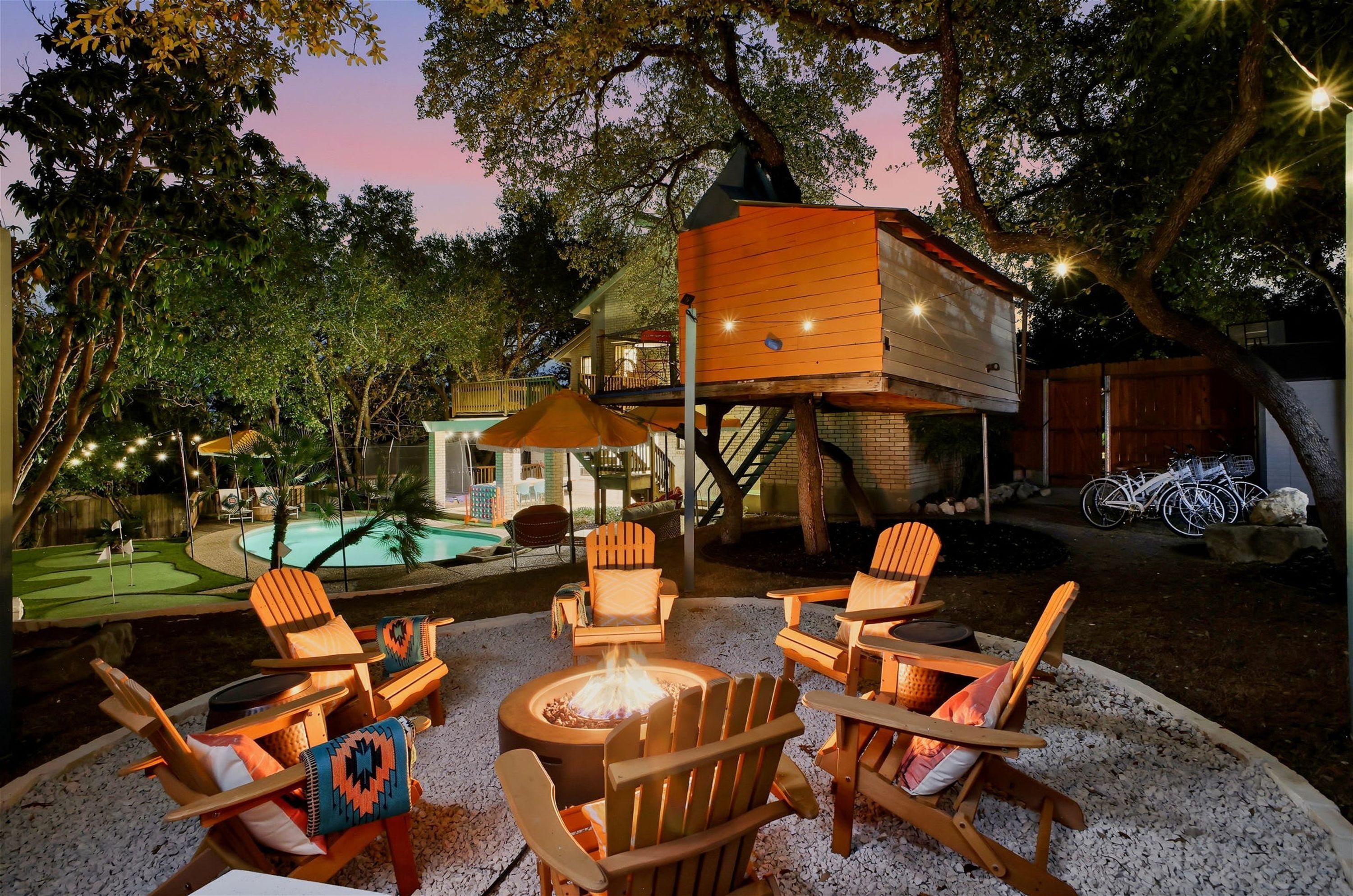 An outdoor seating area with Adirondack chairs and a fire pit, surrounded by trees and a pool.