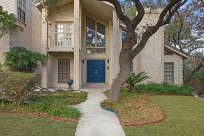 A two-story house with a front entrance, a covered porch, and a large tree in the front yard. - Backyard Paradise - Pool - Tree House - Hot Tub (San Antonio)
