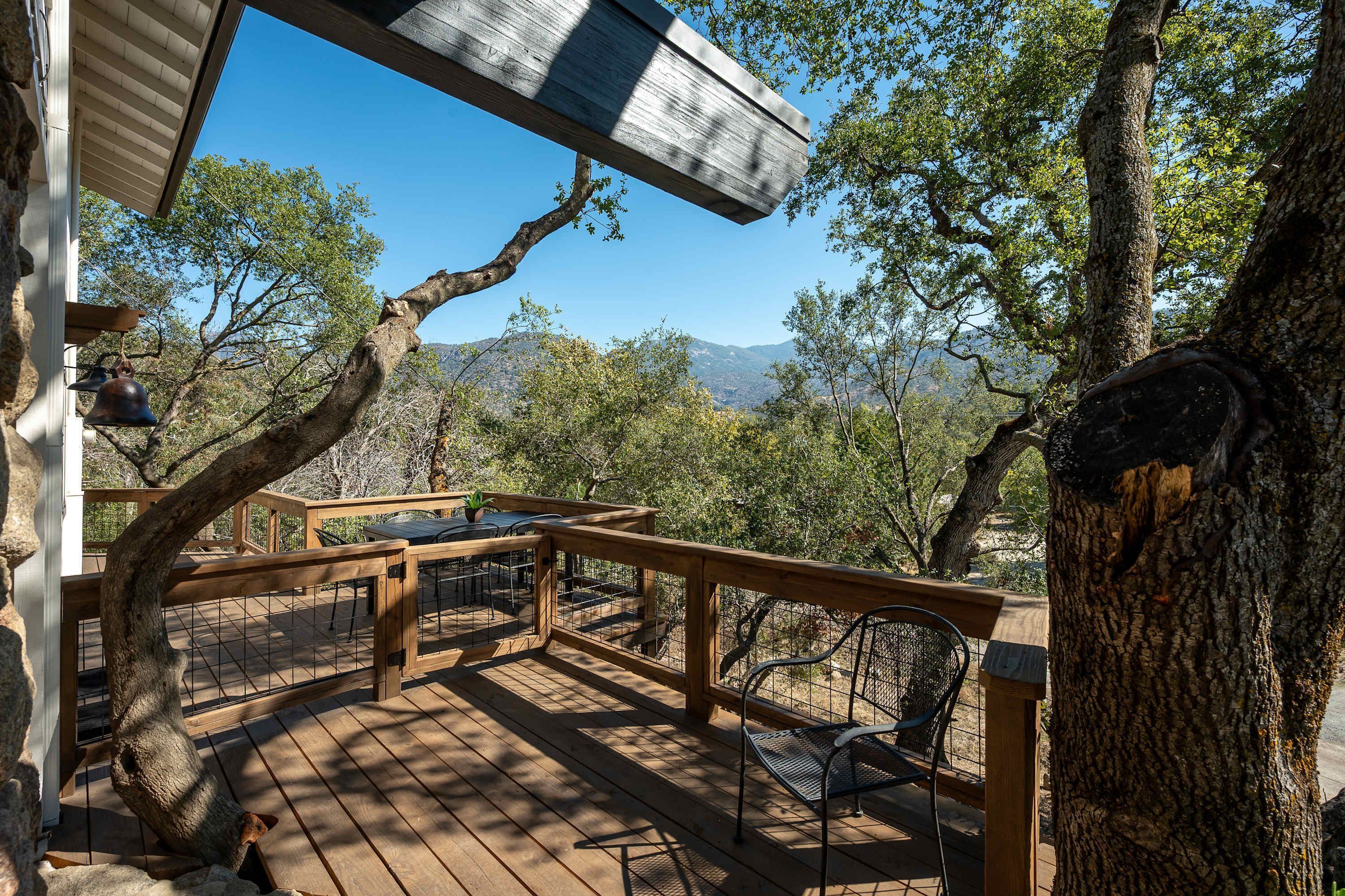 A wooden deck with a table and chairs, surrounded by trees and a mountainous backdrop.
