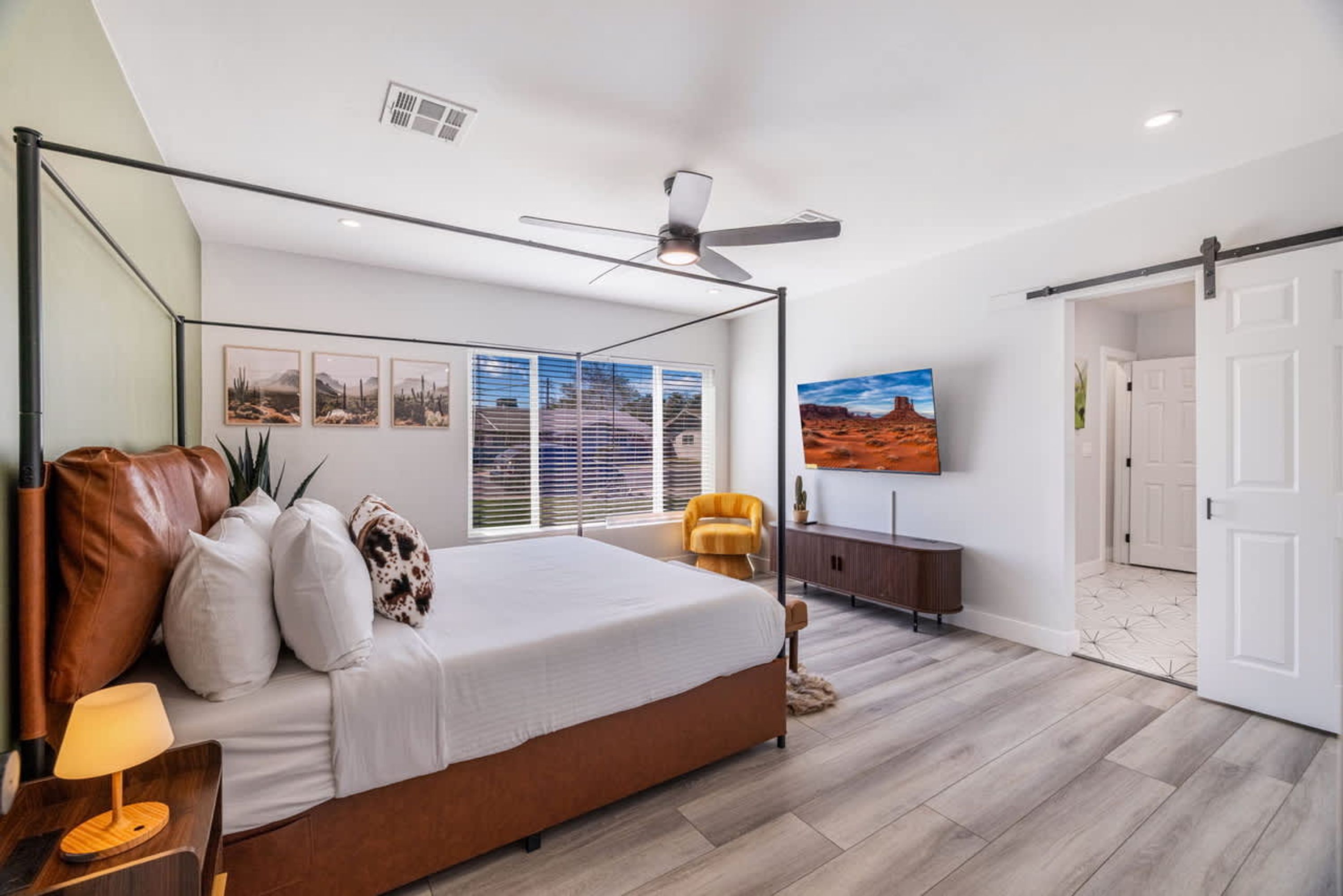 Een moderne slaapkamer met een hemelbed, een plafondventilator, een groot raam met zonwering, een flatscreen televisie en een schuifdeur.