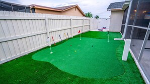 Een achtertuin met kunstgras, een witte schutting en een kleine putting green met vlaggen.