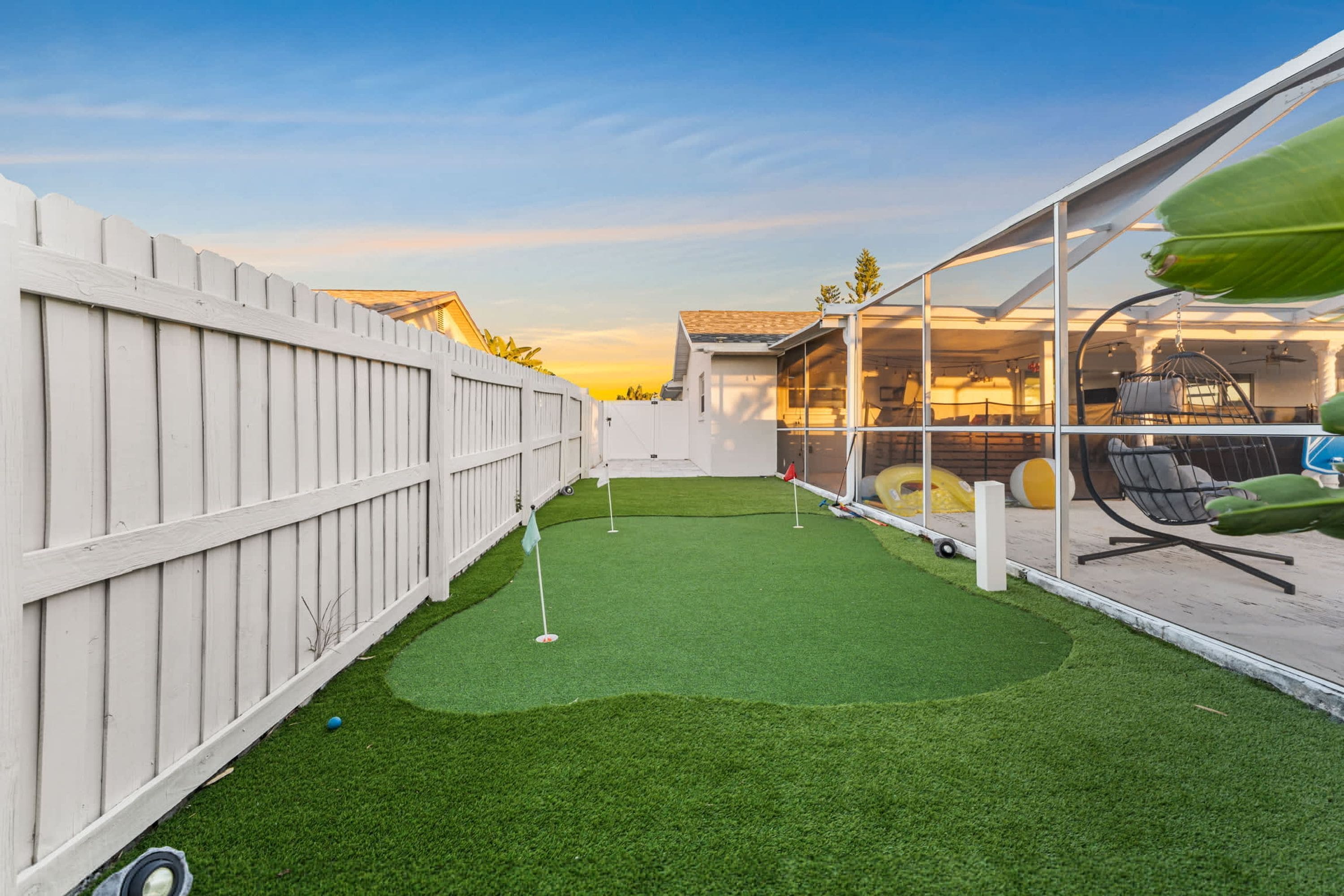 A backyard with artificial grass, a white fence, and a glass enclosure with outdoor furniture.