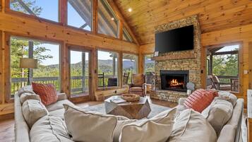 A cozy cabin interior with a stone fireplace, comfortable seating, and large windows offering scenic views.