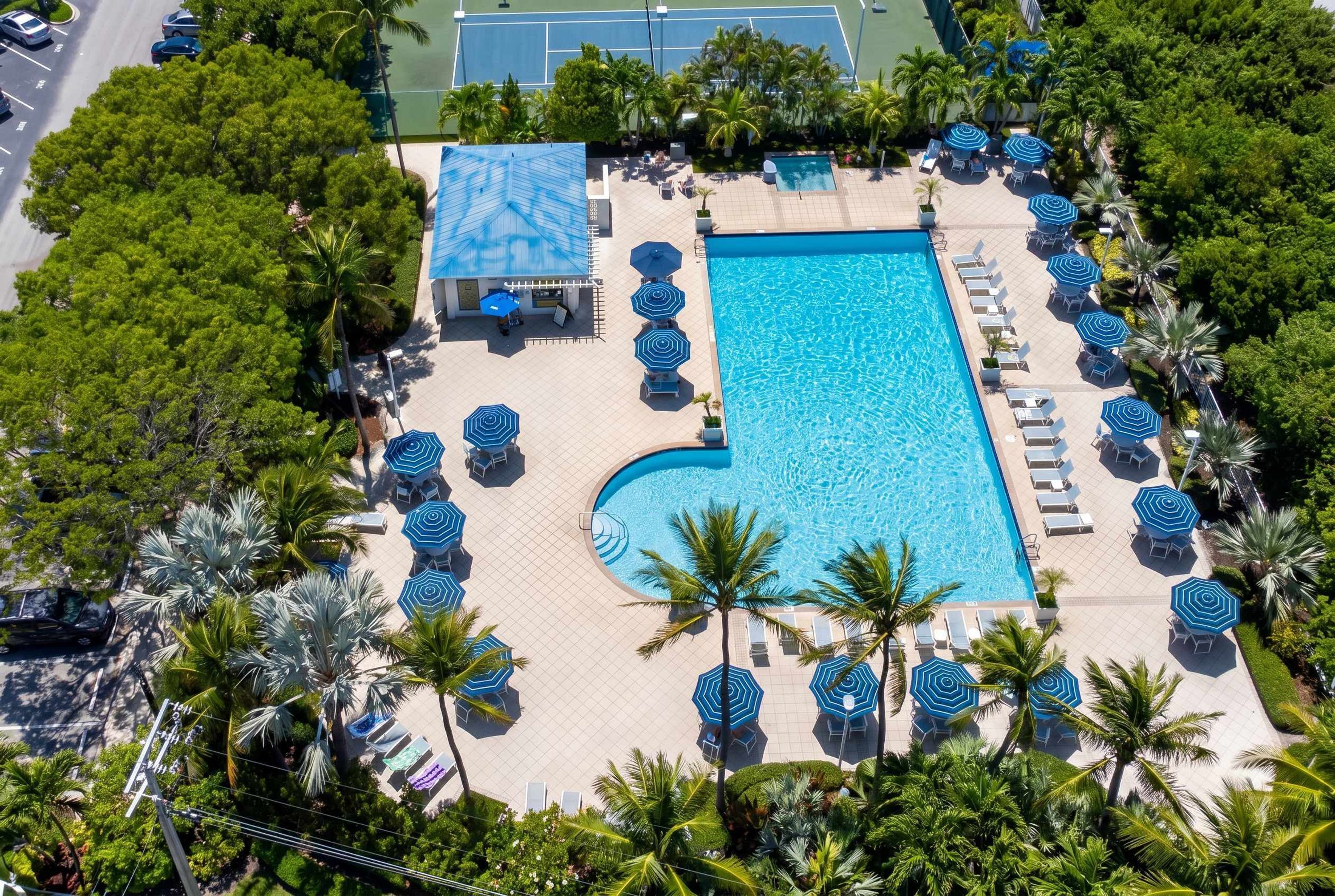 Vista aérea de um resort com uma piscina retangular cercada por espreguiçadeiras e guarda-sóis, palmeiras e uma quadra de tênis ao fundo.
