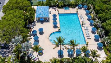 Vista aérea de um resort com uma piscina retangular cercada por espreguiçadeiras e guarda-sóis, palmeiras e uma quadra de tênis ao fundo.