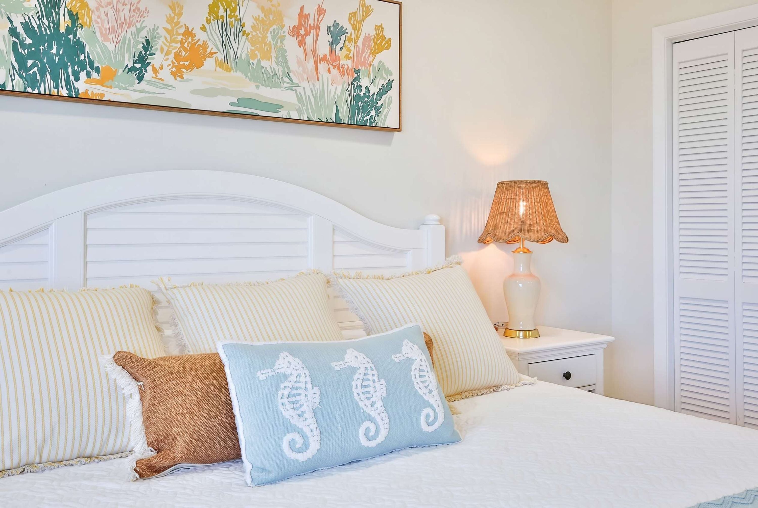 Une chambre à coucher avec un lit blanc, des oreillers à rayures, un coussin en forme d’hippocampe, une table de chevet avec une lampe et une œuvre d’art murale.