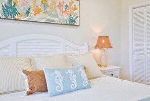 Ein Schlafzimmer mit einem weißen Bett, gestreifter Bettwäsche, einem Kissen in Form eines Seepferdes, einem Nachttisch mit einer Lampe und einem Wandbild.