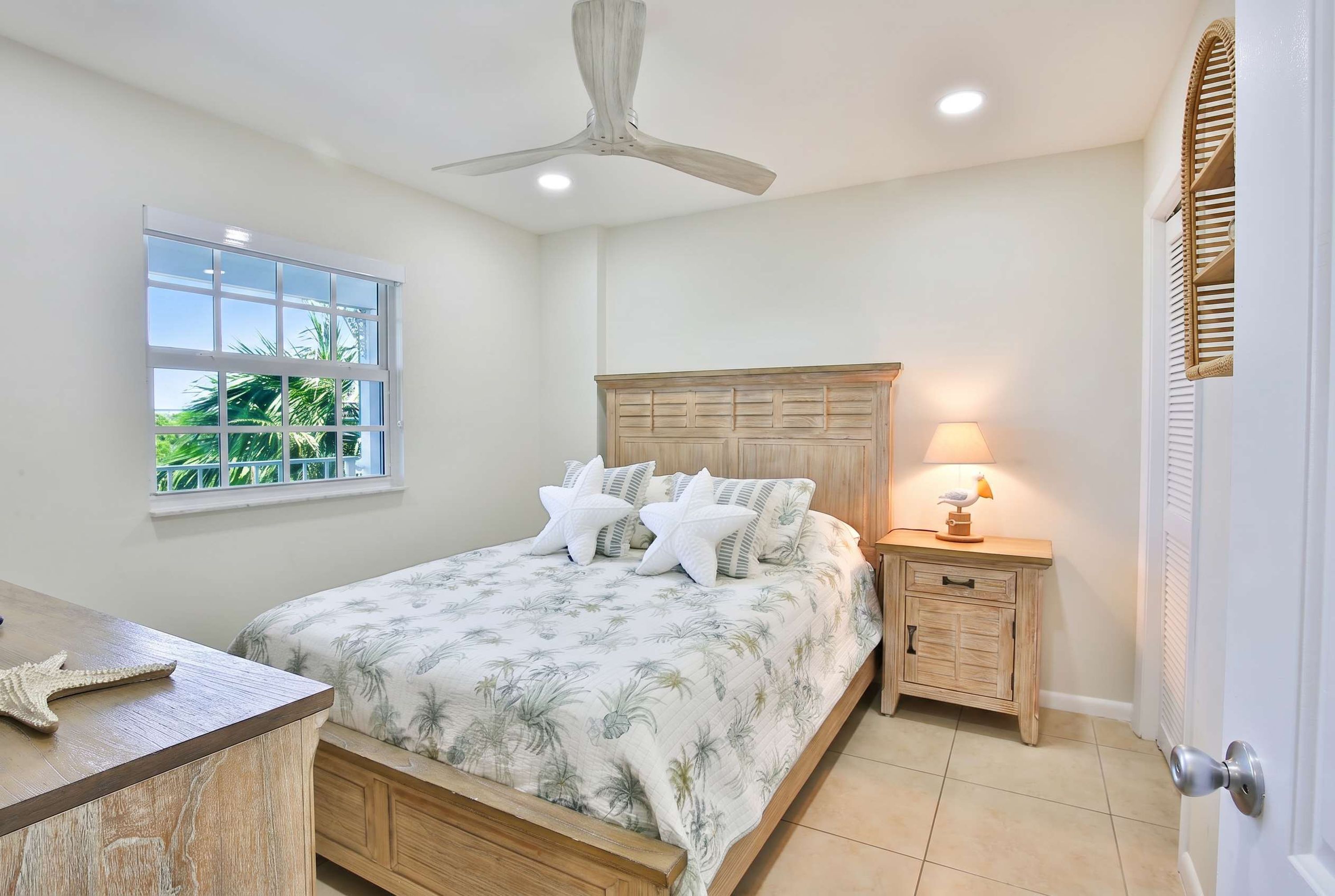 Une chambre à coucher comprenant un lit, une table de chevet, un ventilateur de plafond, une fenêtre donnant sur un espace verdoyant et une porte avec un cadre blanc.