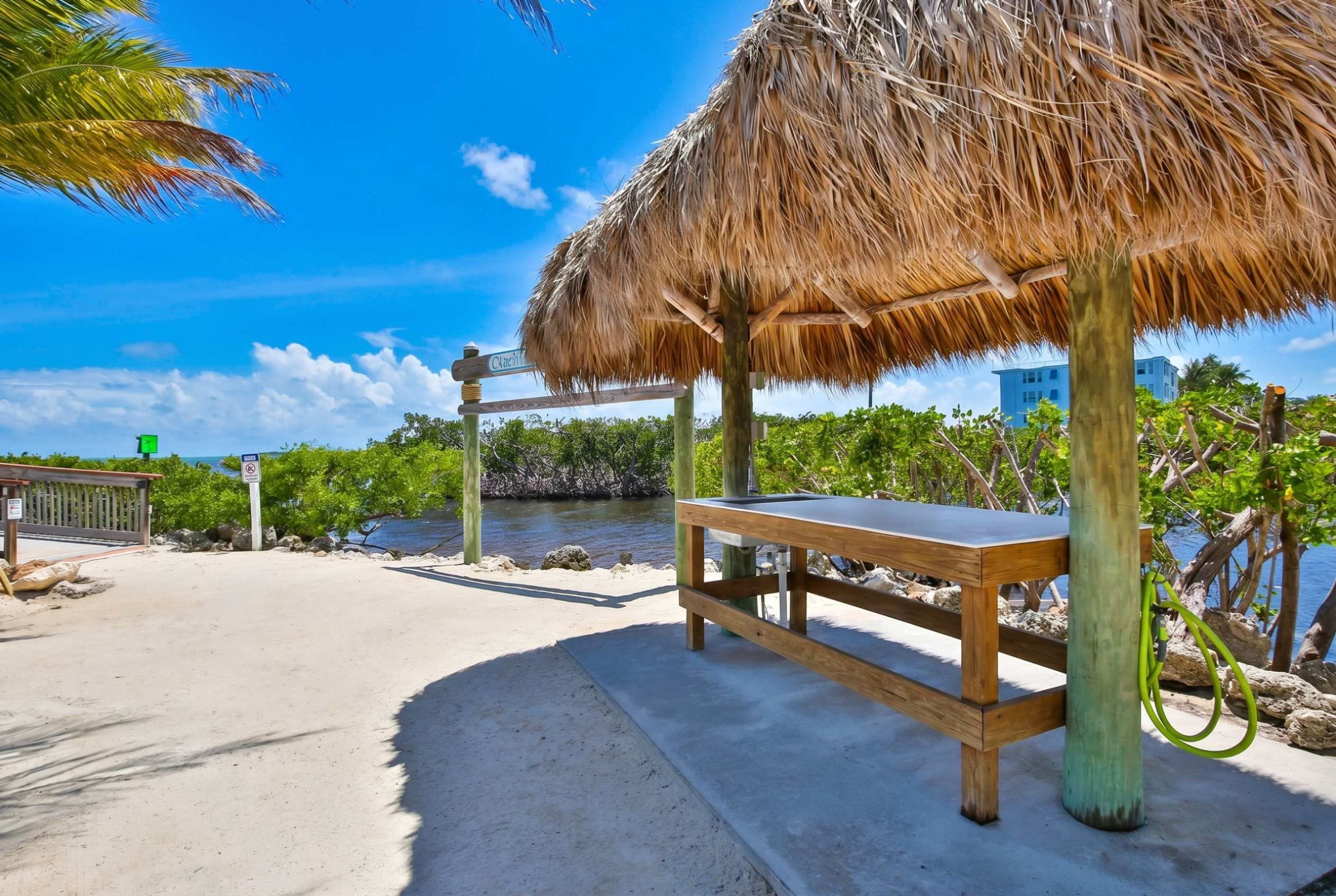 Zona de playa con una estructura de techo de paja, un banco de madera y vista al océano y edificios al fondo.
