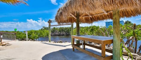 Zona de playa con una estructura de techo de paja, un banco de madera y vista al océano y edificios al fondo.