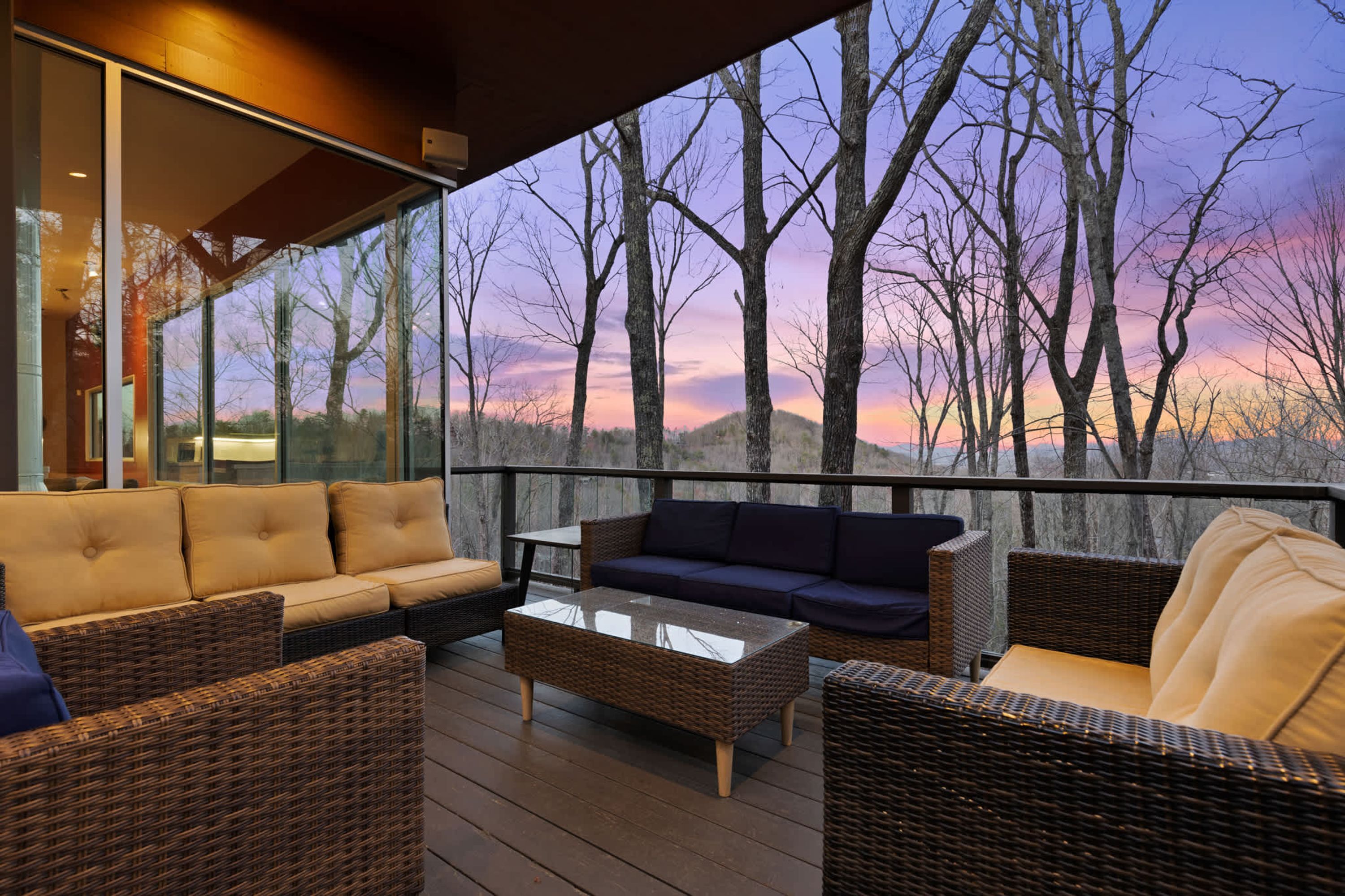 A deck with wicker furniture and a glass coffee table, overlooking a forested landscape.