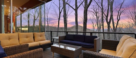A deck with wicker furniture and a glass coffee table, overlooking a forested landscape.