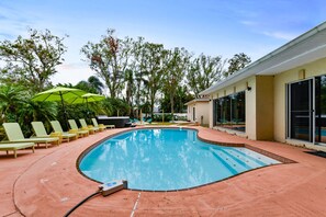 Piscina con sillas de descanso y sombrillas, rodeada de árboles y un edificio con ventanales grandes.
