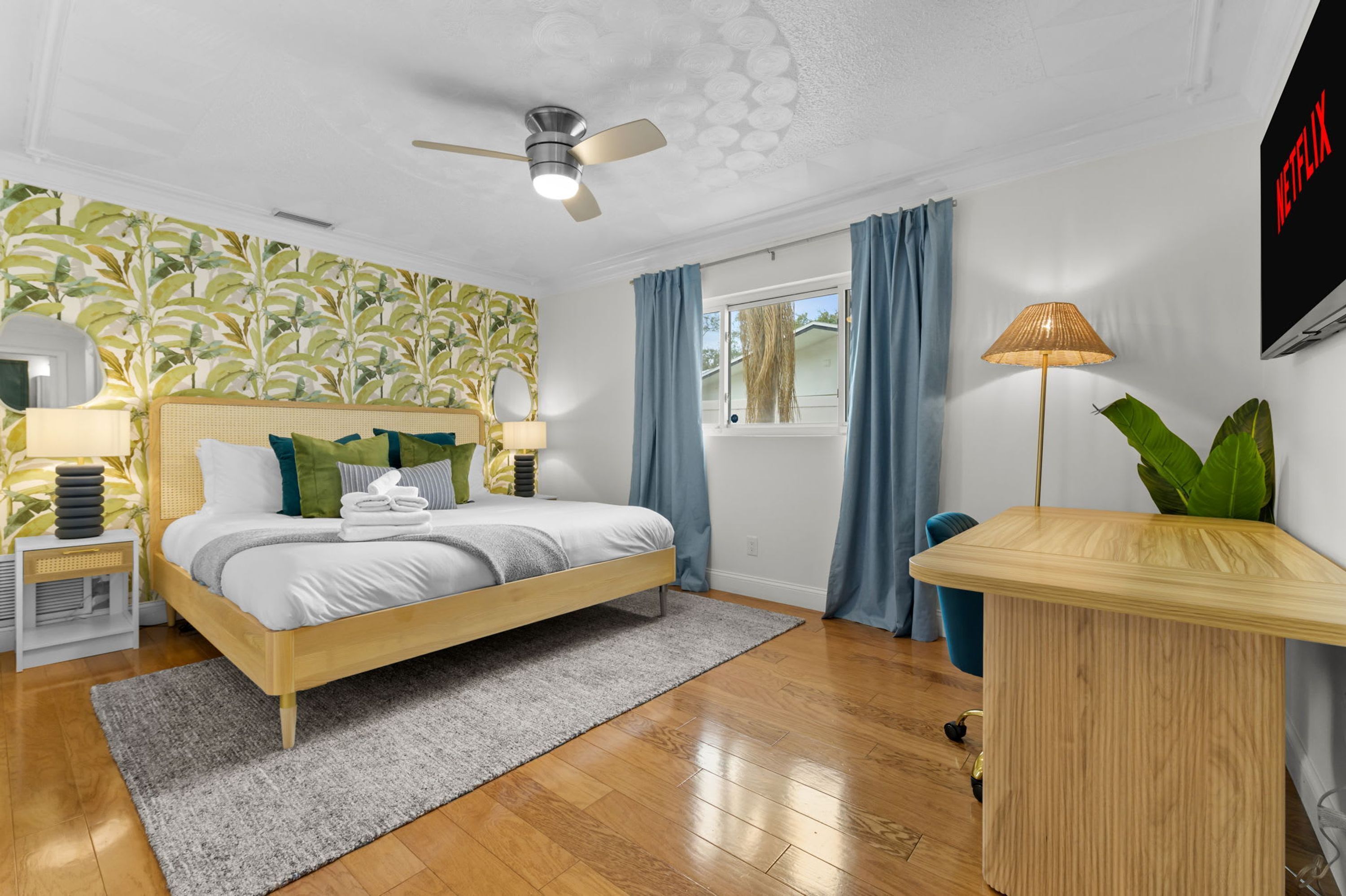 Un dormitorio moderno con cama, ventilador de techo, ventana con cortinas, lámpara de noche y televisor montado en la pared.