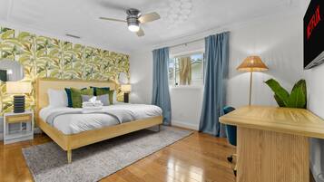 Un dormitorio moderno con cama, ventilador de techo, ventana con cortinas, lámpara de noche y televisor montado en la pared.