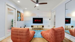 Ein modernes Wohnzimmer mit Deckenventilator, einem großen Flachbildfernseher, der 'NETFLIX' anzeigt, und zwei orangefarbenen Sesseln, die einen blauen Teppich gegenüberstehen.