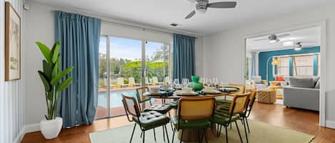 Un coin repas avec une table dressée pour quatre personnes, entourée de chaises, d’un ventilateur de plafond et offrant une vue sur une piscine et la verdure environnante.