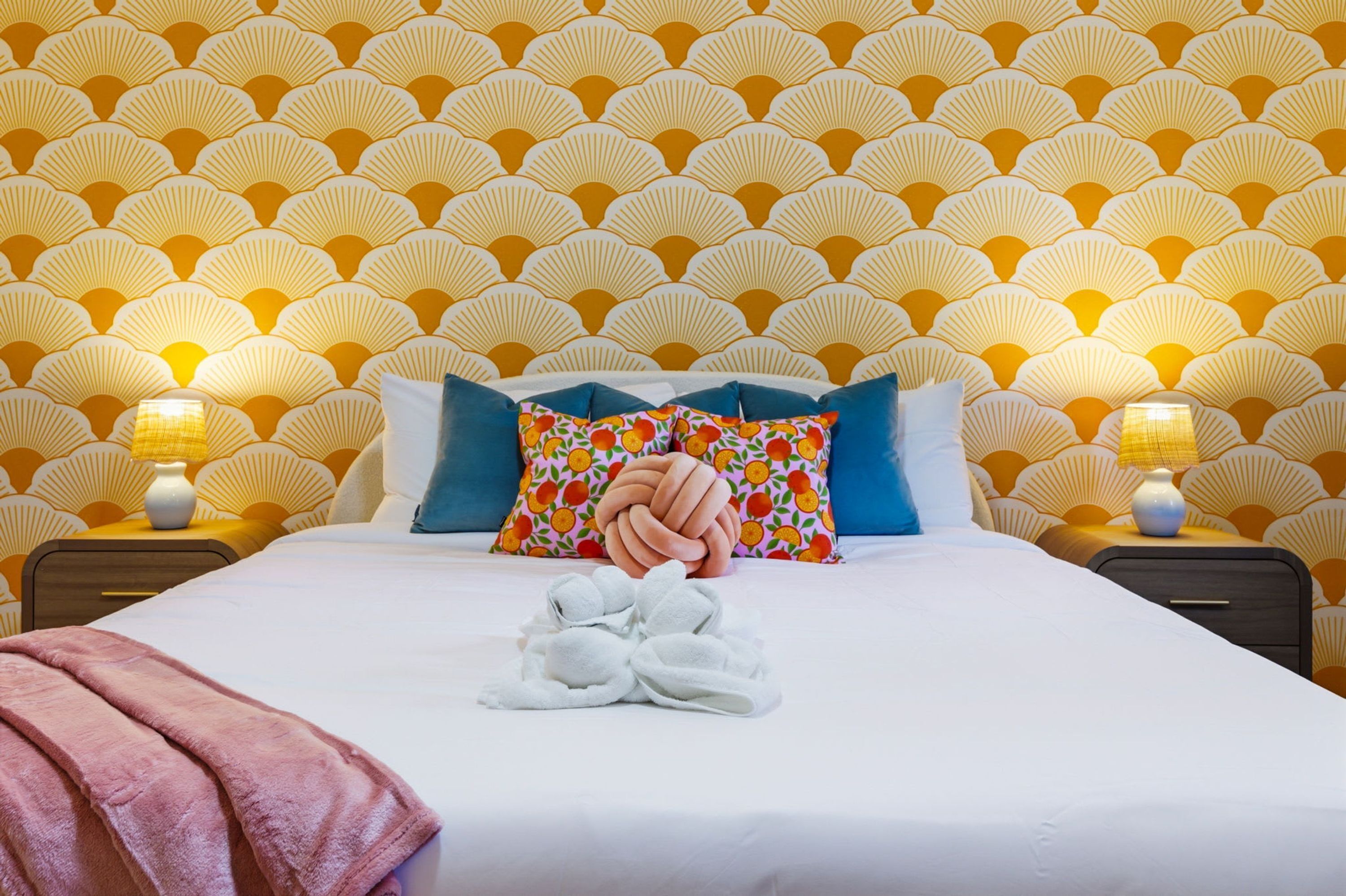 Et hotelværelse med tapet med mønster, en pænt redt seng med et pink håndklæde og to sengeborde med lamper.