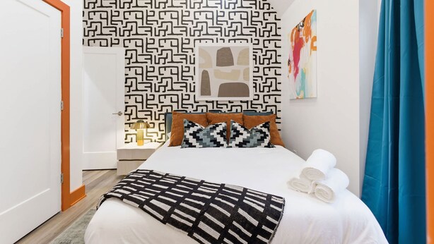 A hotel room with a patterned wall, a bed with white linens, a black and white throw blanket, and a blue curtain. - Victory Lane Villa Game Room - Hot Tub - Firepit (Indianapolis)