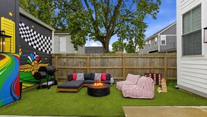 A backyard with a checkered wall mural, a barbecue grill, a fire pit, and a seating area with striped cushions. - Victory Lane Villa Game Room - Hot Tub - Firepit (Indianapolis)