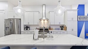 A modern kitchen with white cabinets, a stainless steel refrigerator, a central island with a marble countertop, and pendant lighting. - Victory Lane Villa Game Room - Hot Tub - Firepit (Indianapolis)