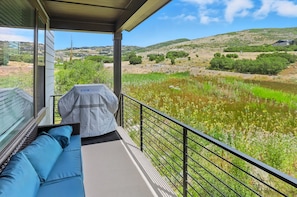 Ein Balkon mit Blick auf einen See und Hügel, einem überdachten Grill und einer blauen Sitzgelegenheit mit Polstern.