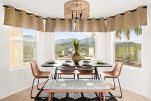 Comedor con una mesa preparada para cuatro, un florero con plantas y vistas a montañas y vegetación.
