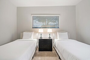 A twin room with two beds, a nightstand with lamps, a window with blinds, and a carpeted floor. - 2 Homes 7BR Sleeps 18 Pool Amazing Mtn Views (Tucson)