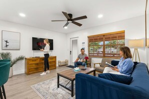 Uma sala de estar com televisão, ventilador de teto, mesa de centro e sofá.