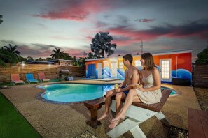 Um casal sentado à beira da piscina com o pôr do sol ao fundo.