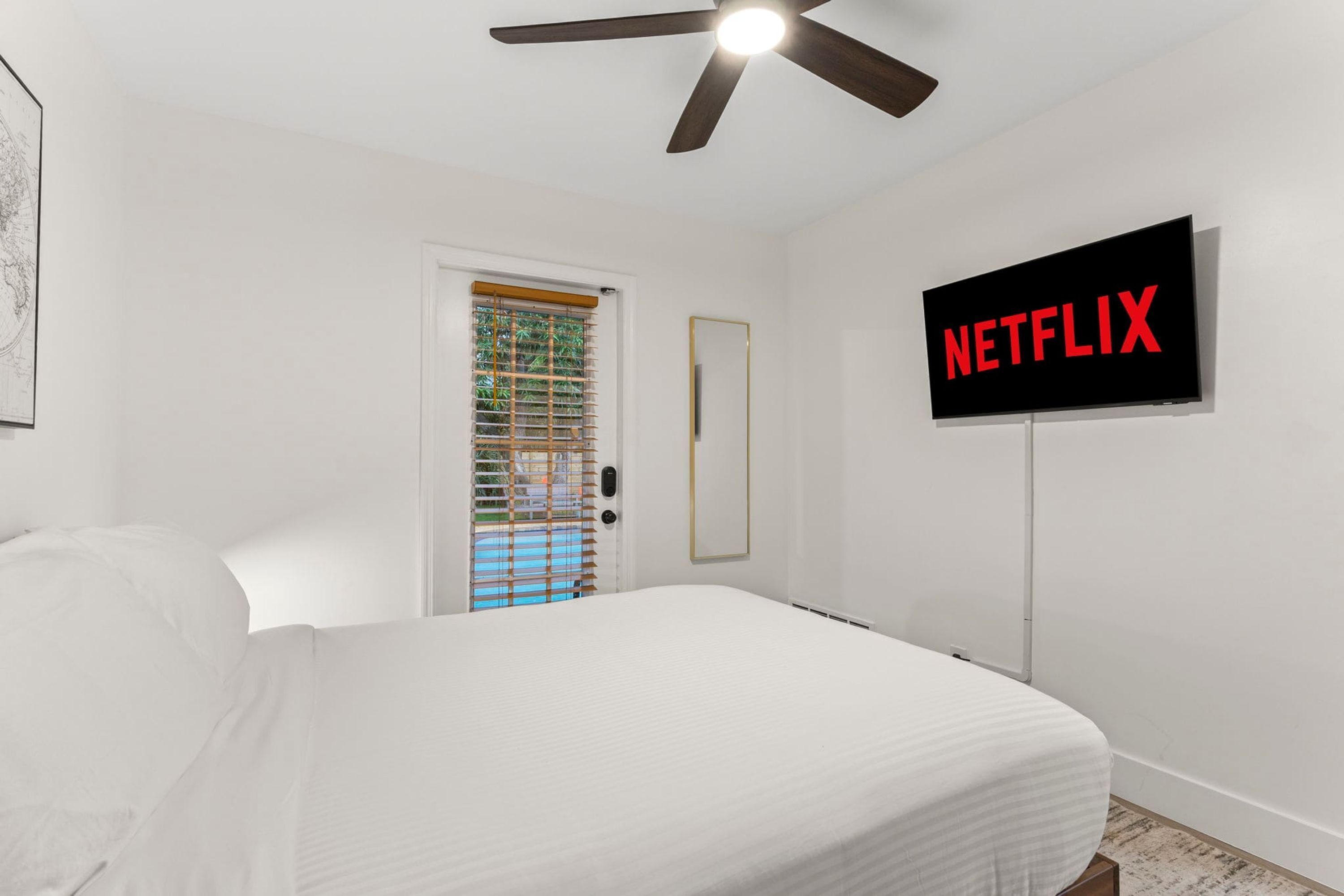 Ein Schlafzimmer mit Bett, einer Tür mit Blick nach draußen, einem Deckenventilator und einem an der Wand befestigten Fernseher, der das Wort 'NETFLIX' anzeigt.
