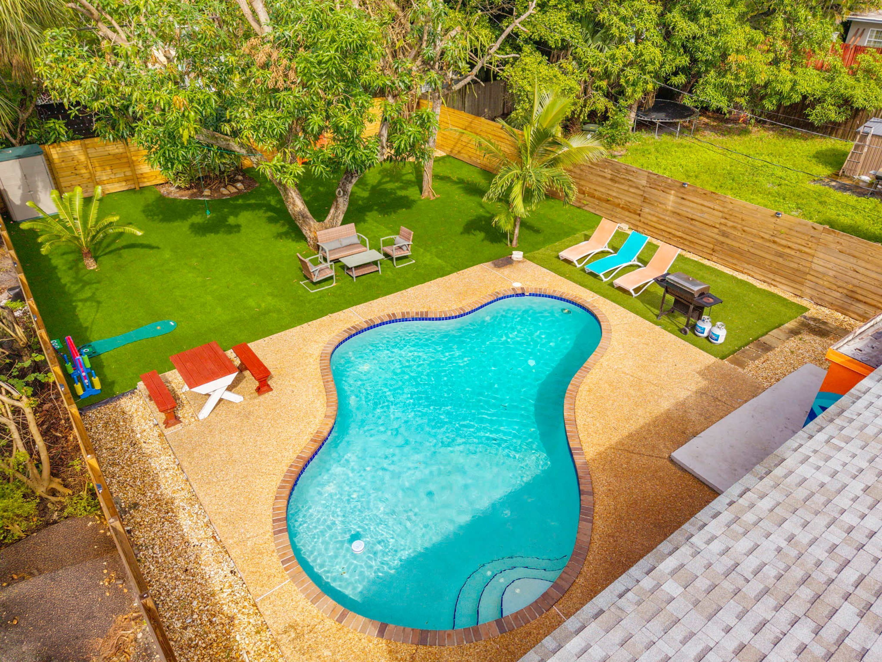 Une piscine dont le bord est revêtu de carrelage, entourée d’une pelouse avec des arbres et une clôture en bois.