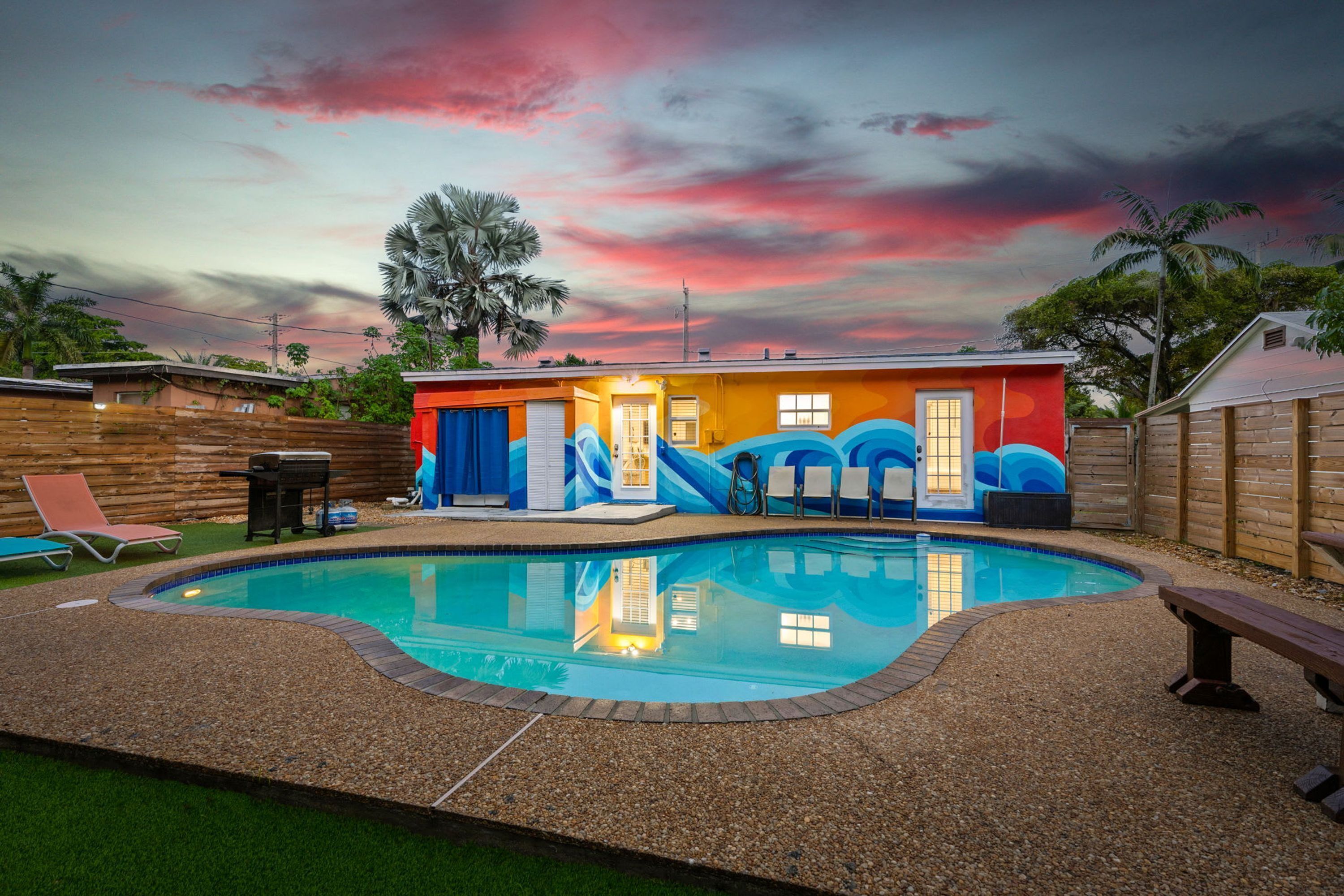 Une piscine avec un toboggan, entourée d’une clôture en bois, et une maison aux murs extérieurs aux couleurs vives.