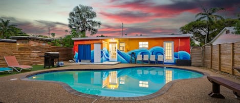 Une piscine avec un toboggan, entourée d’une clôture en bois, et une maison aux murs extérieurs aux couleurs vives.