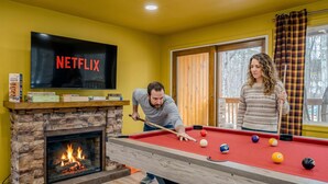 Una habitación con mesa de billar, chimenea, televisor mostrando 'NETFLIX' y ventana con cortinas.